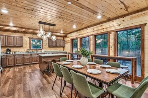 A spacious kitchen with wooden cabinets, a dining area with a large table and chairs, and large windows offering a view of the forest. - Huge Cabin 22 Beds | Indoor Pool+ Movie Theater (Sevierville)