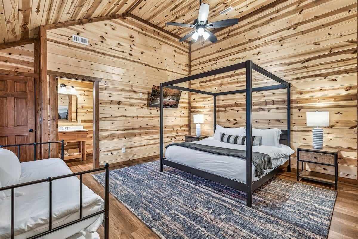 A four-poster bed in a wooden cabin room with a ceiling fan, a nightstand, and a lamp.