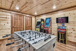 A room with wooden paneling, a foosball table, and arcade game machines. - Huge Cabin 22 Beds | Indoor Pool+ Movie Theater (Sevierville)