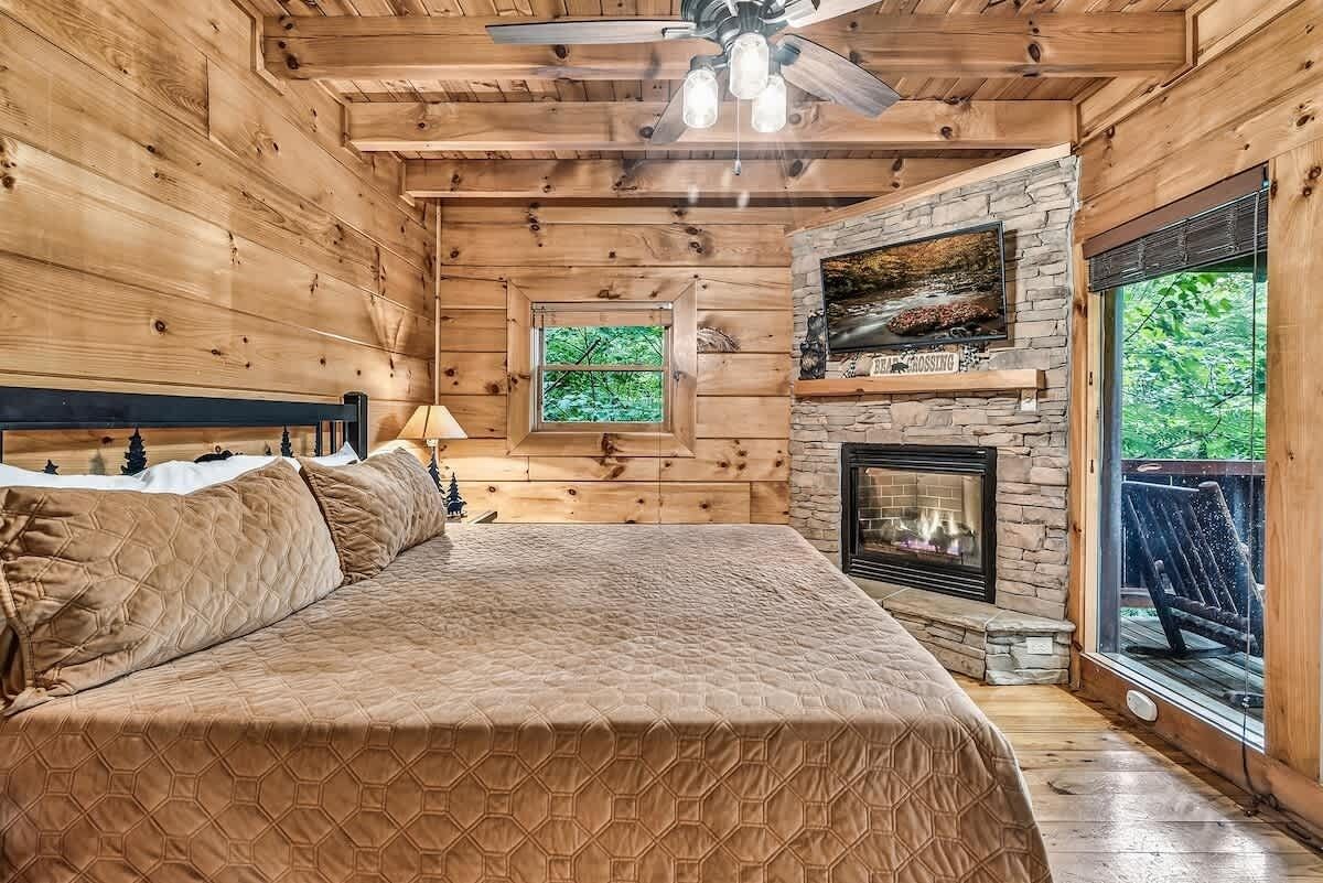 Une chambre à coucher située dans un chalet en rondins, avec un lit, une cheminée en pierre, un ventilateur de plafond et une fenêtre donnant sur la verdure.