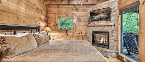 Een slaapkamer in een blokhut met een bed, een stenen openhaard, een plafondventilator en een raam met uitzicht op groen.