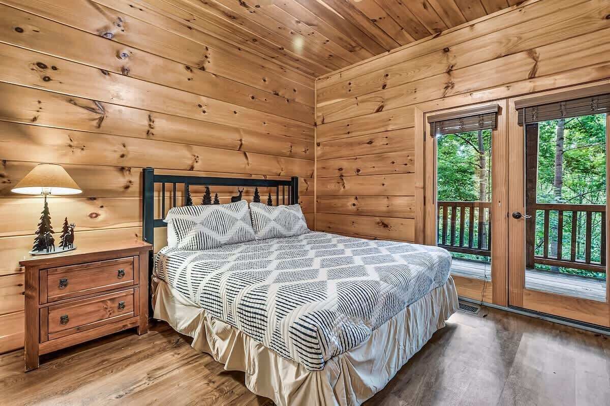Ein Schlafzimmer einer Holzhütte mit Bett, Nachttisch, Kommode und einem großen Fenster mit Blick auf Bäume.