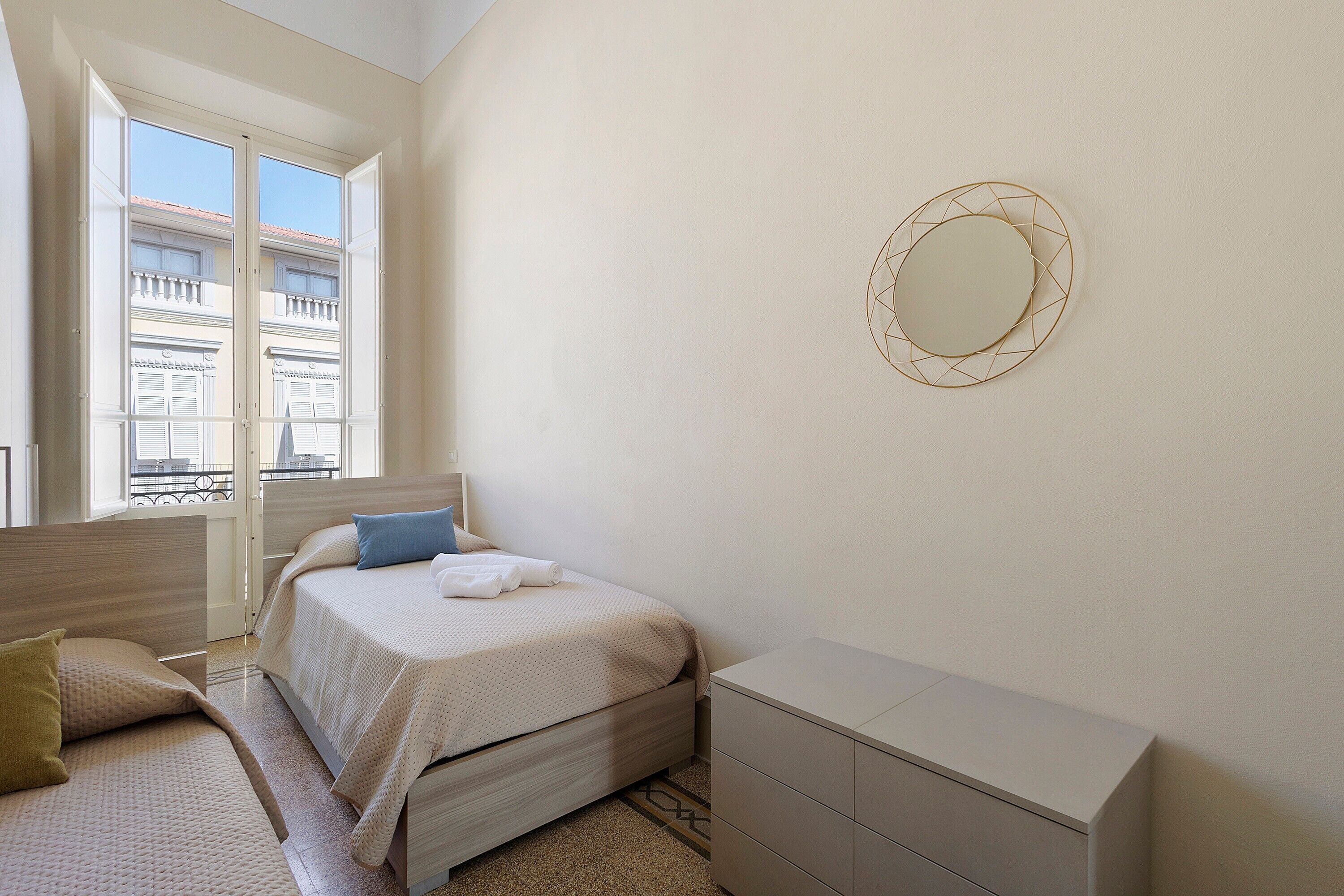 Une chambre à coucher avec un lit, une commode, un miroir et une fenêtre donnant sur des immeubles.