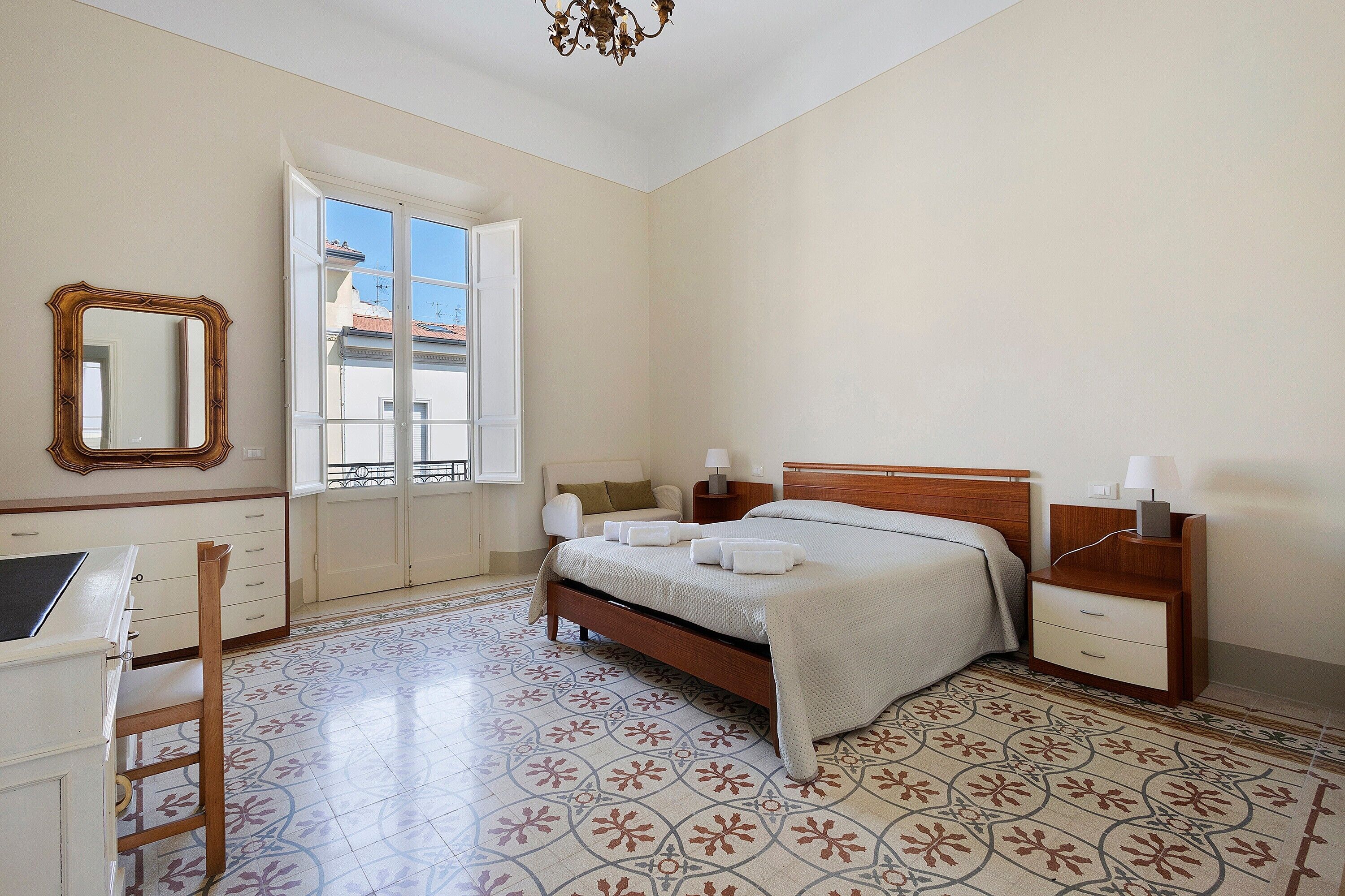 Une chambre à coucher comprenant un lit, des tables de chevet, une chaise, un bureau, un miroir et une fenêtre donnant sur des immeubles.