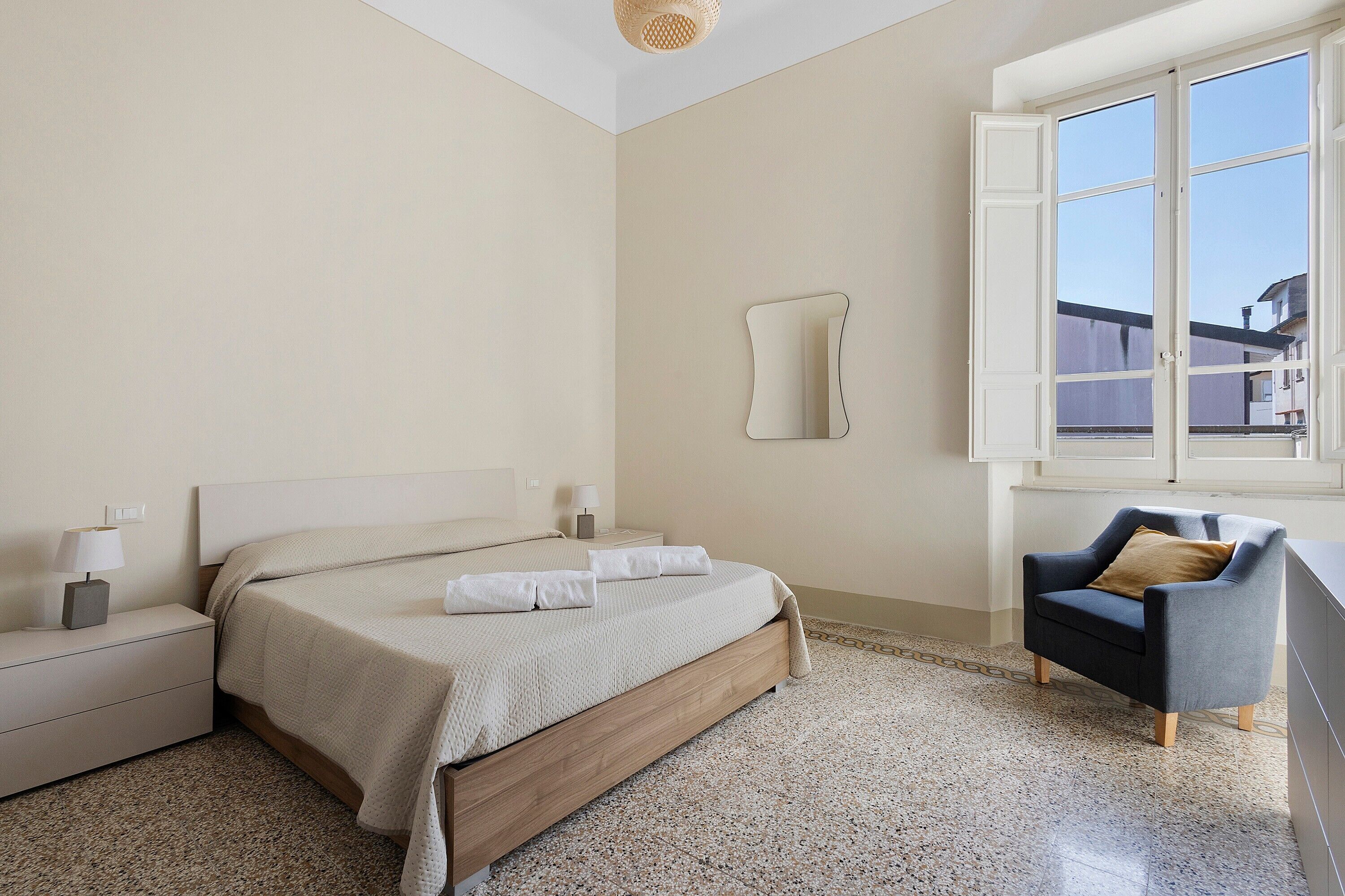 Una camera da letto con un letto, comodini, una sedia, una finestra e uno specchio fissato alla parete.