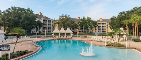 A large swimming pool with a fountain, surrounded by lounge chairs and a building with multiple balconies.