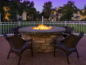 A circular stone fire pit with seating around it, surrounded by a white fence, with buildings in the background. - Sheraton Vistana Resort 2 Bedroom Villa (Orlando)