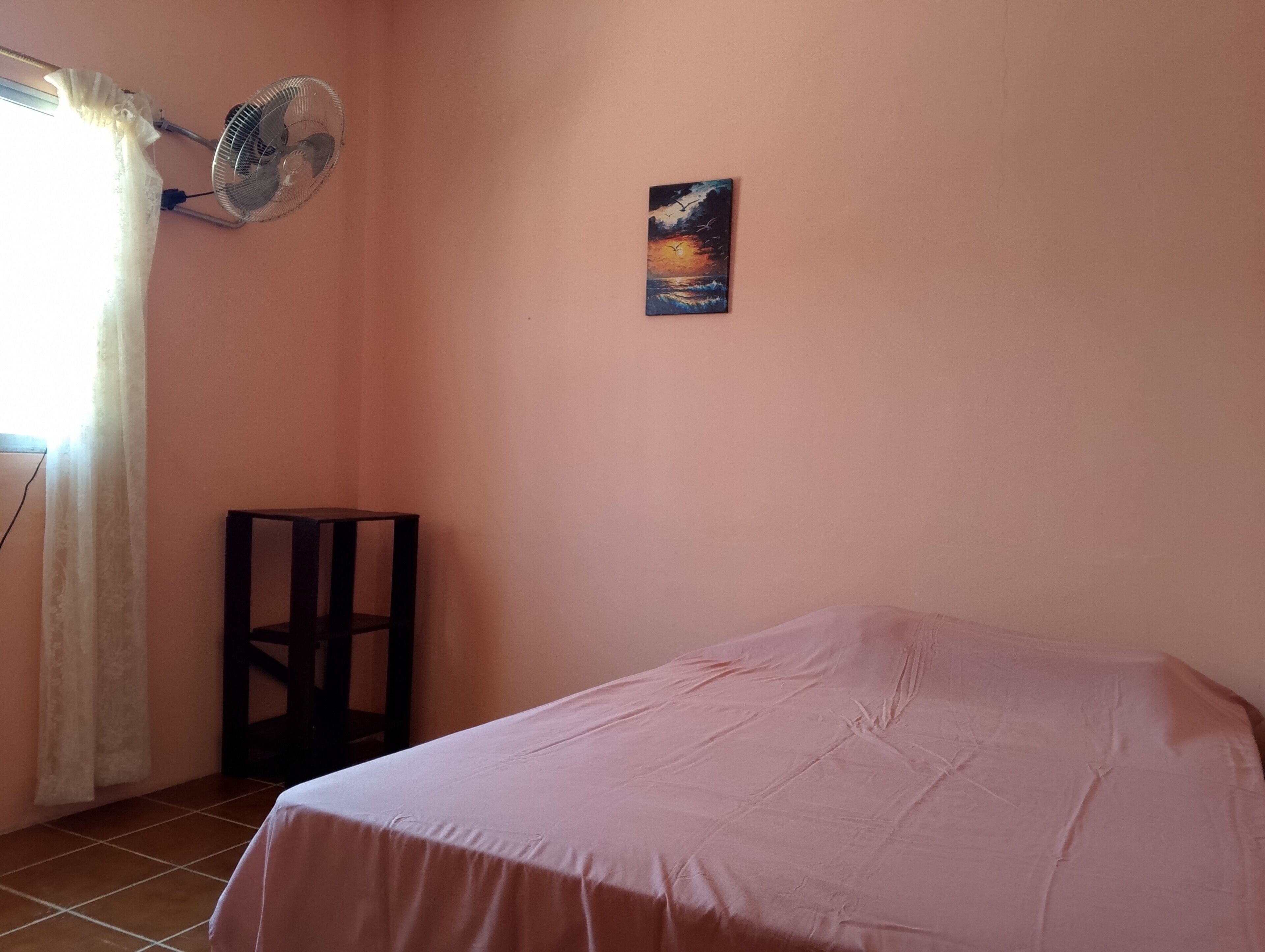 A bedroom with a bed, a standing fan, a painting, and a window with curtains.