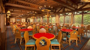 A spacious dining area with multiple tables and chairs, featuring a rustic wooden ceiling and large windows allowing natural light.