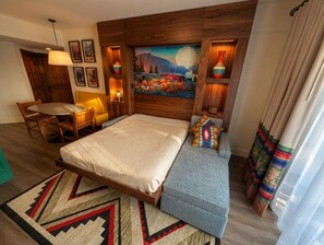 Una habitación de hotel con una cama, zona de estar, un cuadro en la pared y una ventana con cortinas.