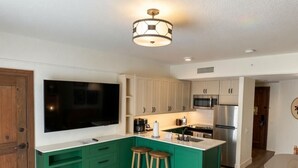 A modern kitchen with green cabinetry, a large flat-screen TV, a stainless steel refrigerator, and a bar stool. - Orlando | Couples Lodge Retreat | Pool + Hot Tub (Lake Buena Vista)