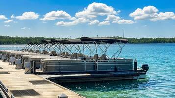 A dock with several pontoon boats equipped with canopies, a motorboat, and a clear view of the water and distant shoreline.