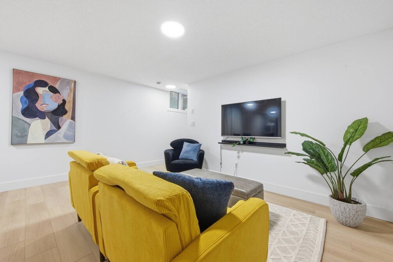 A living room with a yellow sofa, a blue armchair, a flat-screen TV, a painting, and a potted plant.