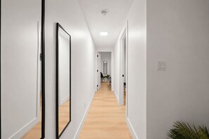 A hallway with white walls and wooden flooring, leading to a room with a door at the end.