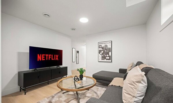 Una sala de estar moderna con un televisor de pantalla plana mostrando 'NETFLIX', un mueble de entretenimiento negro, una mesa de centro redonda de vidrio, un sofá gris y una obra de arte abstracto enmarcada en la pared.