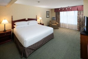 Ein Hotelzimmer mit einem großen Bett, zwei Nachttischlampen, einem Sessel, einem Fernseher und einem Fenster mit Vorhängen.