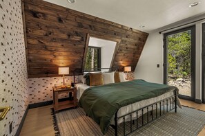 A bedroom with a sloped ceiling, a large bed, two bedside lamps, a nightstand, and a view of greenery through a sliding door.