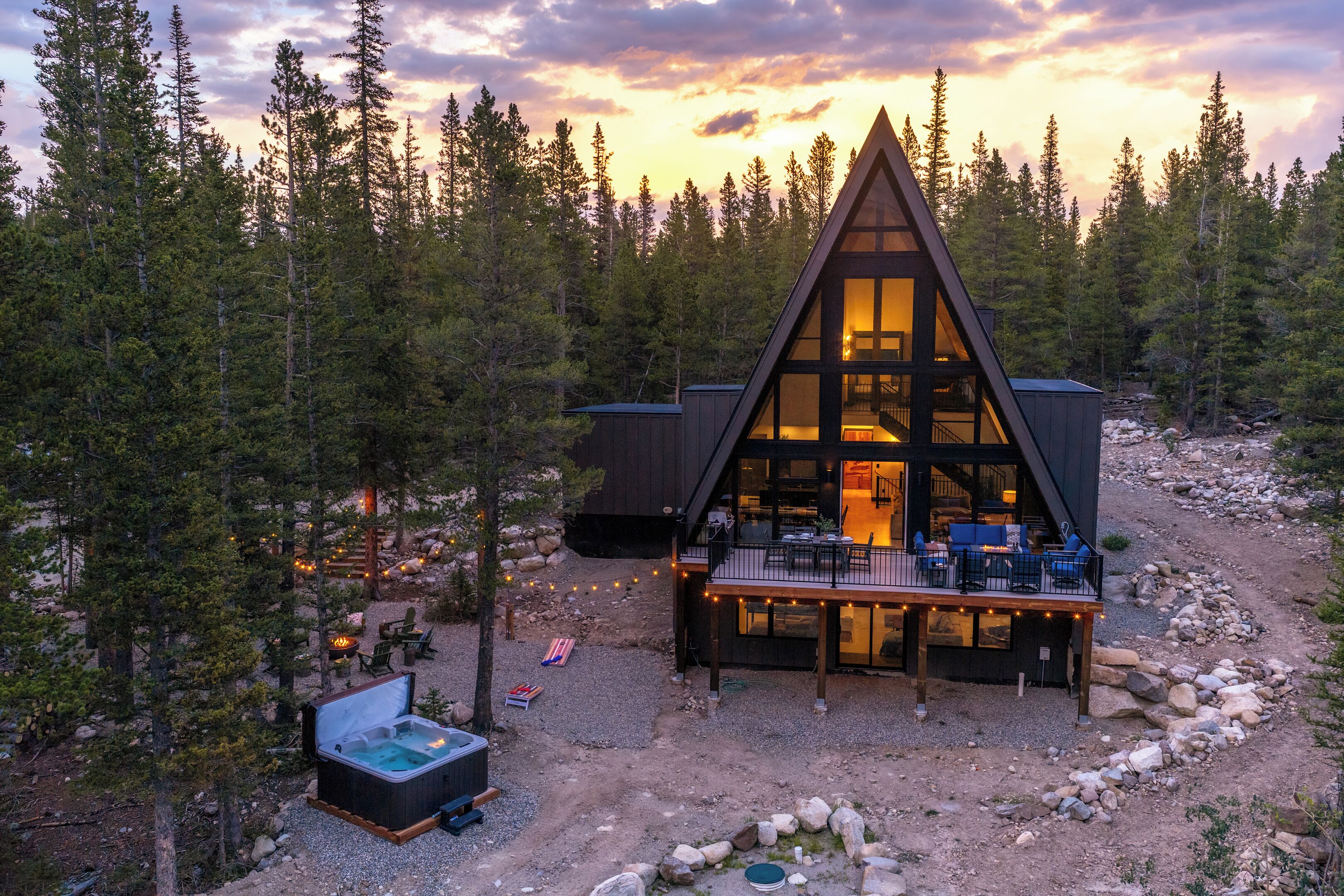 Eine Hütte mit Satteldach, umgeben von einem Wald, mit einer Terrasse, Sitzgelegenheiten im Freien und einem Whirlpool.