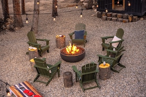 A fire pit surrounded by Adirondack chairs, with a gravel ground and string lights overhead.