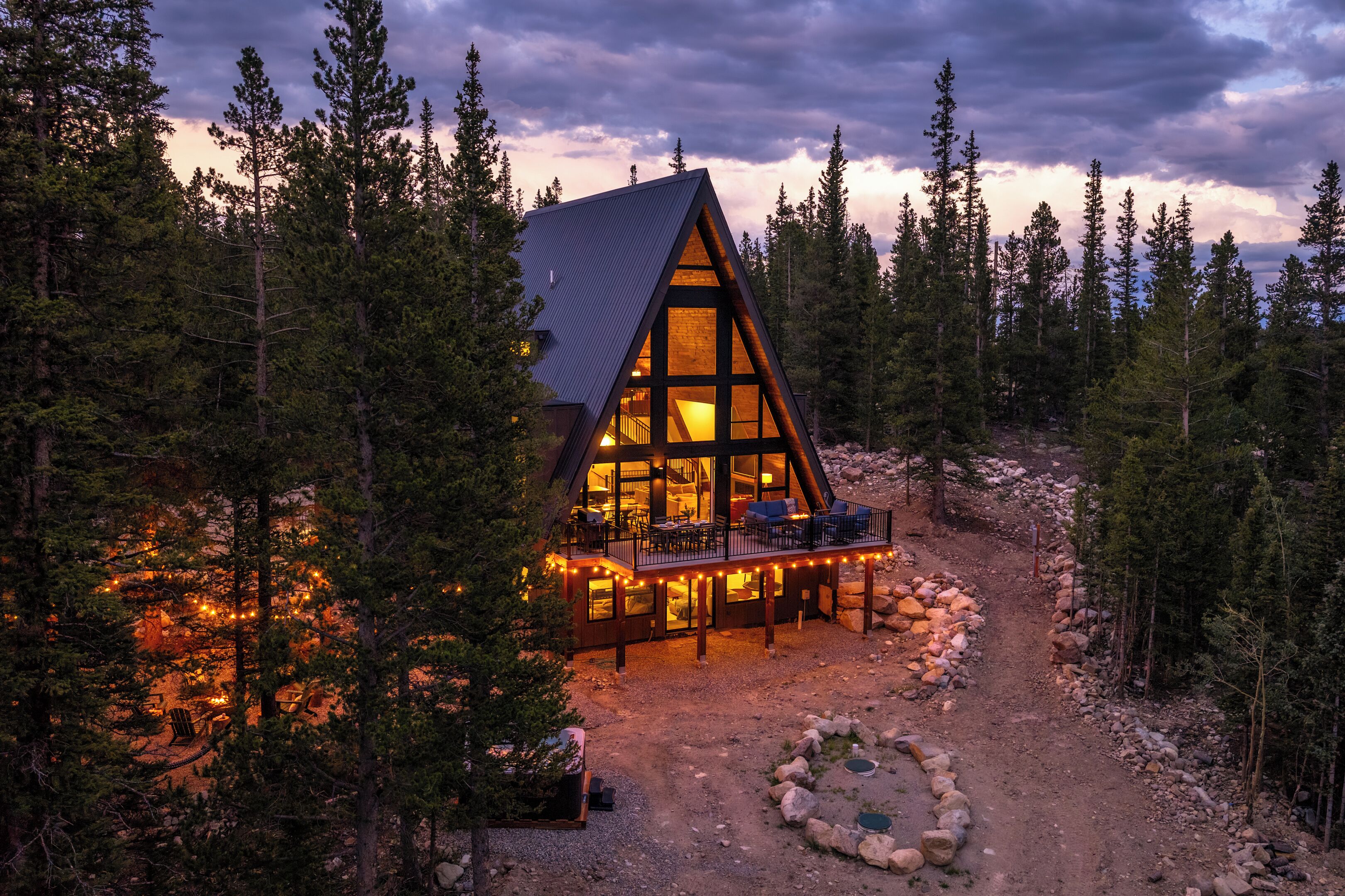 Une cabane dotée d’un toit triangulaire, entourée d’arbres, et d’une terrasse avec des sièges d’extérieur.