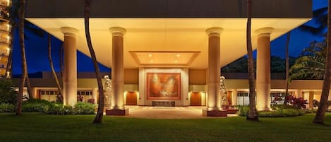 Fachada de hotel con una gran obra de arte central, palmeras y buena iluminación.