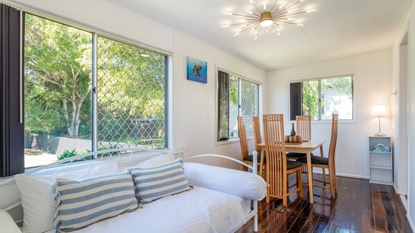 Une pièce avec un canapé blanc, une table à manger avec des chaises, de grandes fenêtres et un luminaire au plafond.