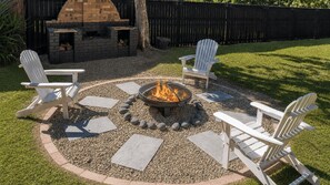 Un patio trasero con un brasero rodeado de sillas Adirondack, una chimenea de ladrillo y una valla de madera.