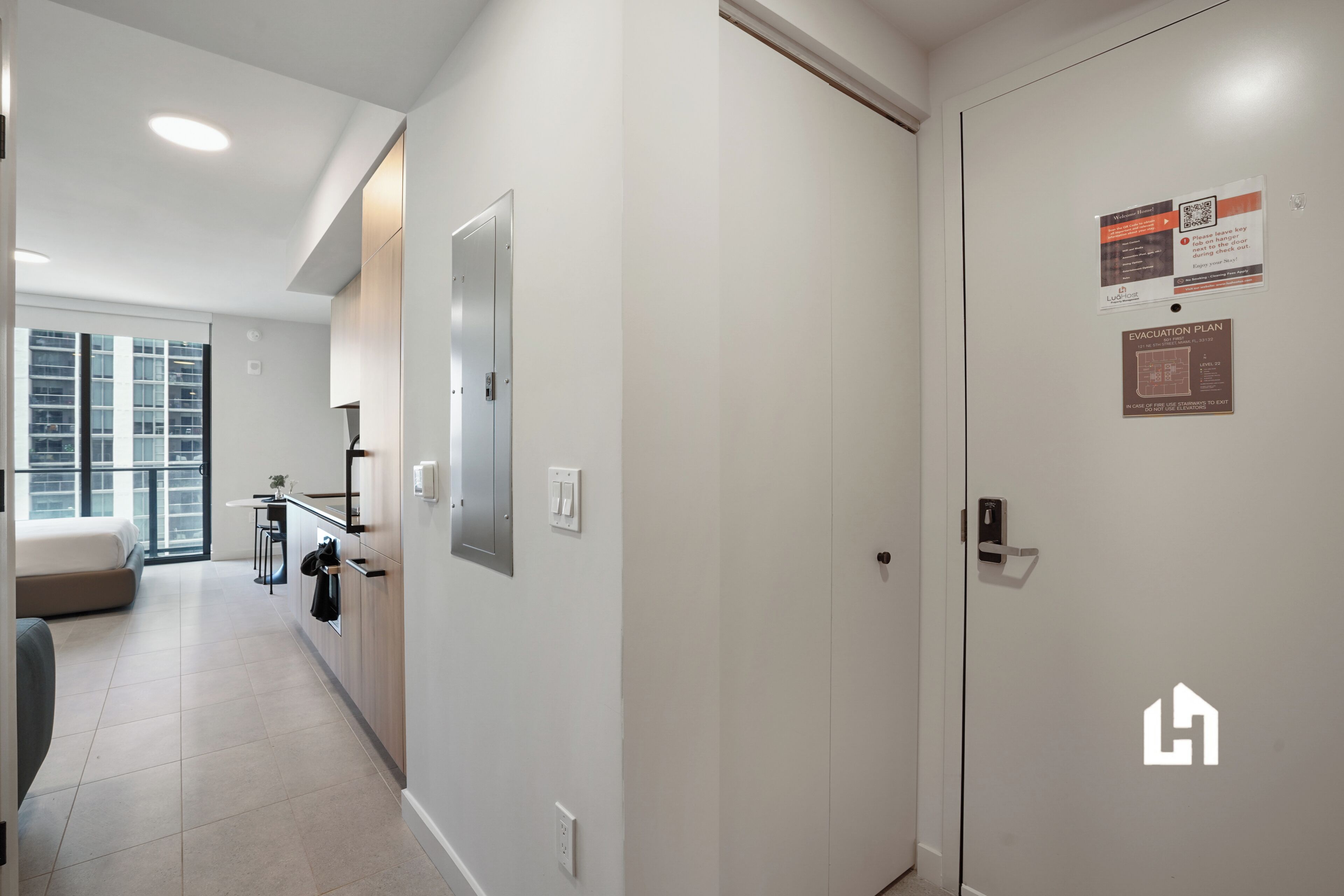A hotel corridor with a closed door, a light switch, and a wall-mounted emergency exit sign.