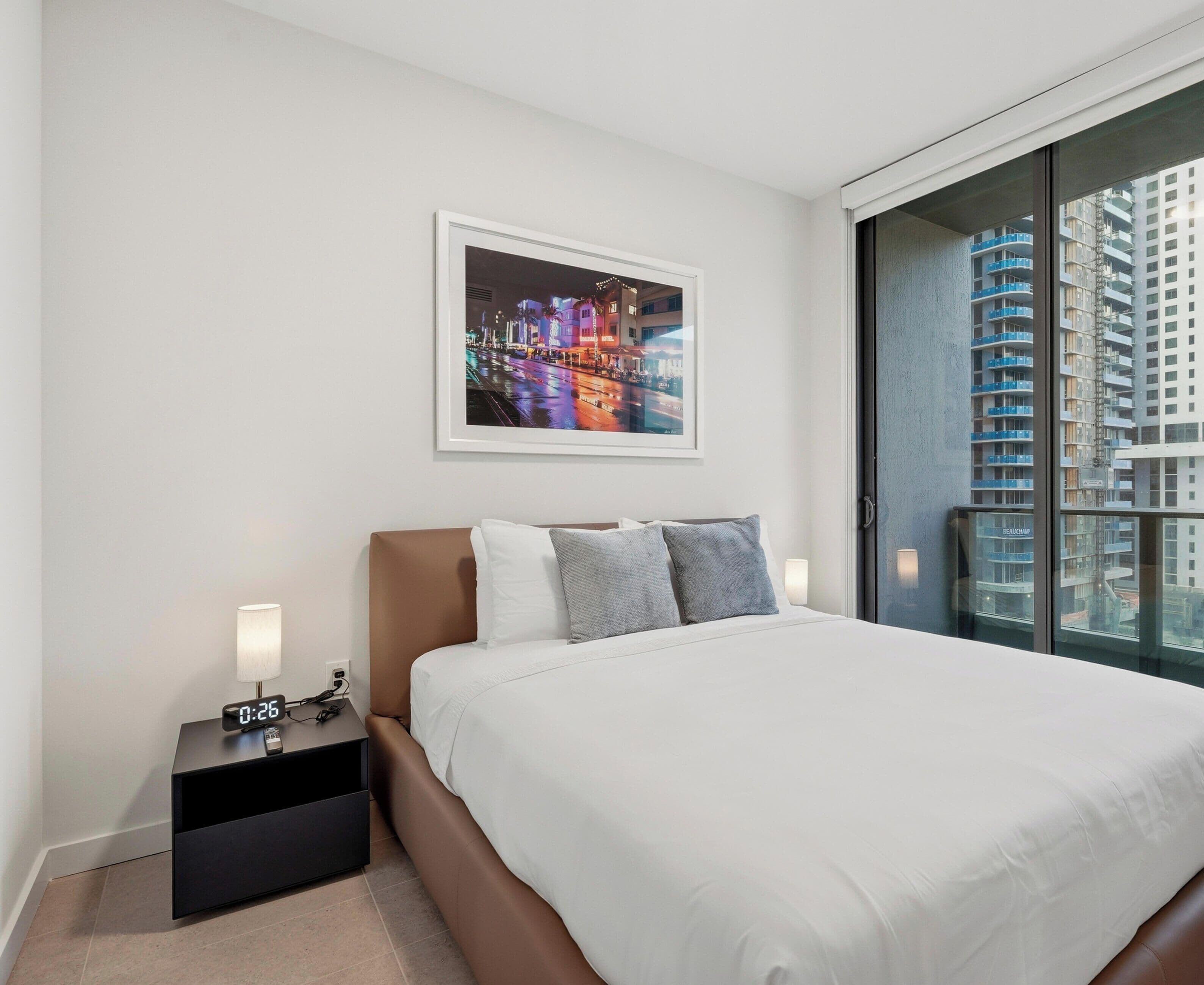 Una habitación de hotel moderna con una cama grande, una mesita de noche con un reloj digital, un cuadro con una vista urbana en la pared y vistas de rascacielos a través de un ventanal amplio.