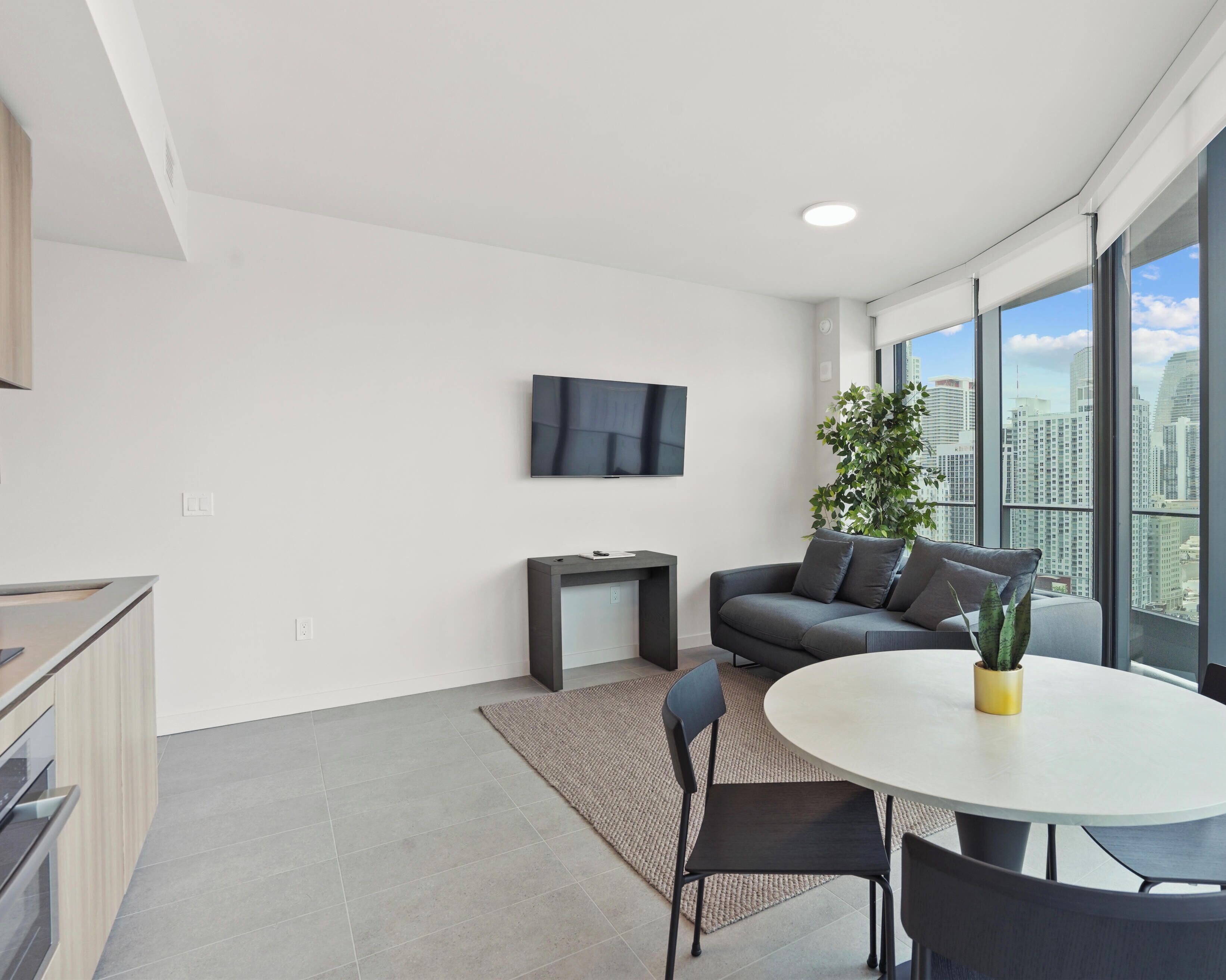 A modern living room with a flat-screen TV, a grey sofa, a round dining table, and a potted plant.
