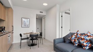 Ein modernes Wohnzimmer mit einem grauen Sofa, bunten Kissen, einem Holztisch und einem Essbereich mit rundem Tisch und Stühlen.