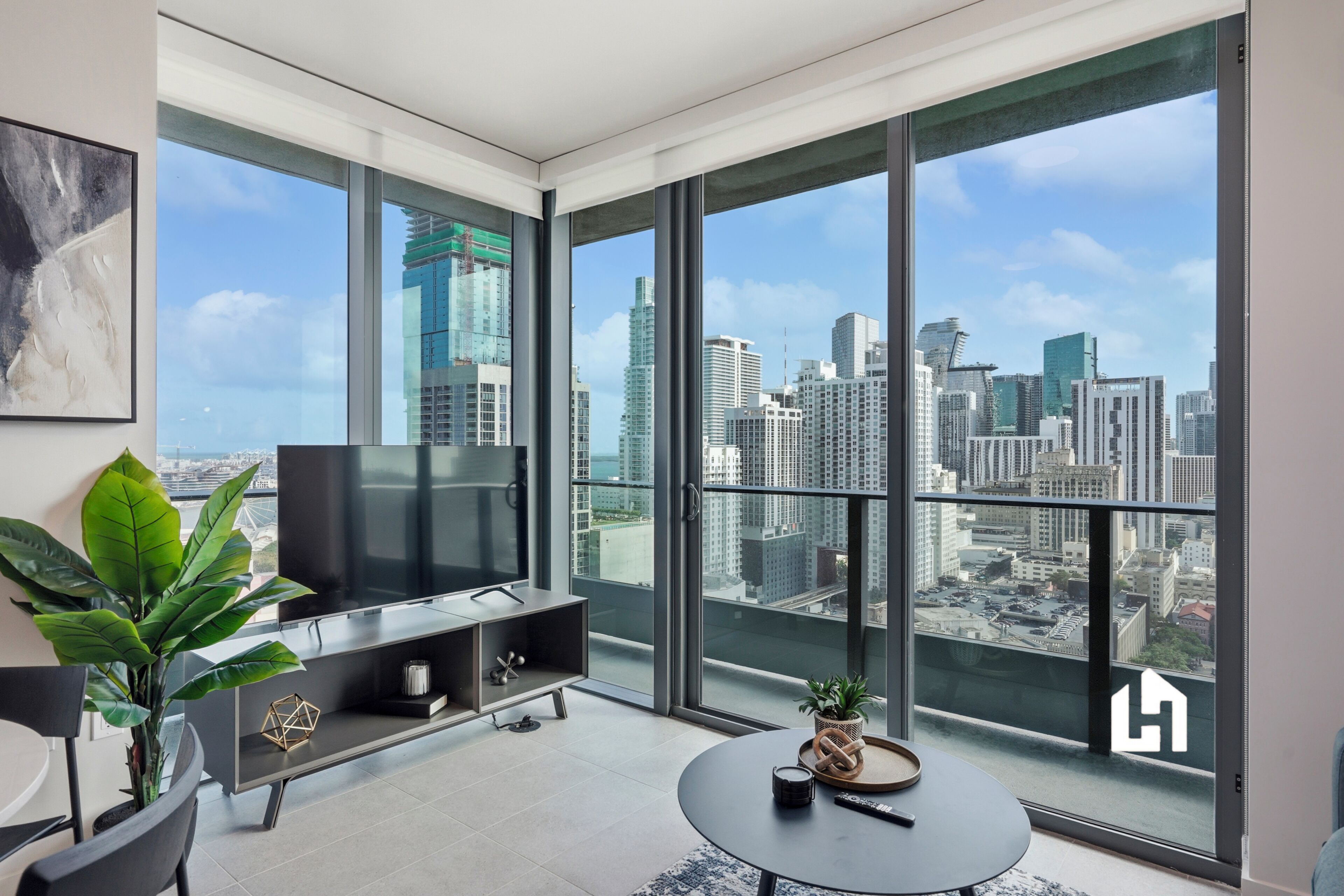 Uma sala de estar moderna com uma televisão grande, uma mesa de centro e vista para o horizonte urbano através de janelas do chão ao teto.