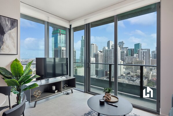 A modern living room with a large TV, a coffee table, and a view of a city skyline through floor-to-ceiling windows.