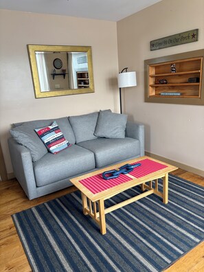 A living room with a grey sofa, a wooden coffee table with a red and blue striped table runner, a blue floor lamp, and a mirror with a gold frame. - Charming 2-bedroom waterfront cottage in Nelson perfect for a relaxing getaway (Nelson)