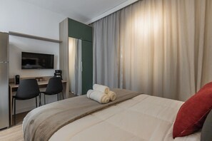 Ein Hotelzimmer mit einem Bett, einem Schreibtisch, zwei Stühlen, einem Fernseher und einem großen Fenster mit Vorhängen.