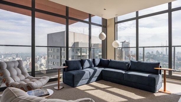 Un salon moderne avec un canapé d’angle, un fauteuil à capitons et une petite table d’appoint. De grandes fenêtres offrent une vue sur la ville.