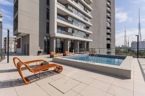 Área de piscina no terraço com espreguiçadeiras e um edifício moderno ao fundo.