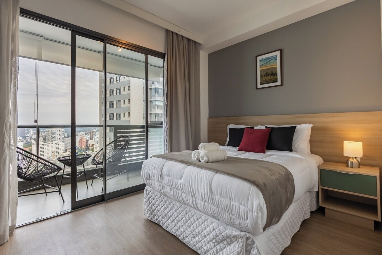 A hotel room with a large bed, a nightstand, a lamp, a chair, and a view of the city through floor-to-ceiling windows.
