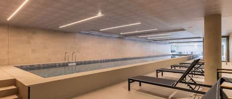 A modern indoor swimming pool with a long bench area and a glass partition.