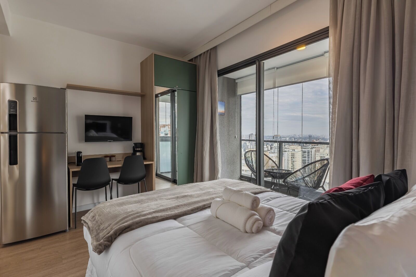 Ett hotellrum med en säng, ett skrivbord, en tv och en balkong med utsikt över staden.