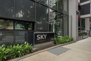 Ein modernes Gebäude mit großer Glasfassade, einem Schild mit der Aufschrift 'SKY Pinheiros 180 Residencial' und einem kleinen Gartenbereich mit Pflanzen.