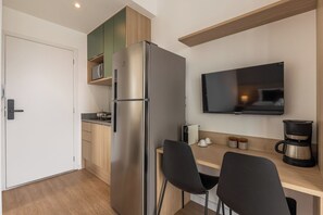 Uma cozinha compacta com geladeira de aço inoxidável, TV de tela plana na parede, cafeteira e duas cadeiras pretas em uma mesa de madeira.