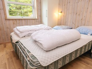 A room with two beds, each with a quilted mattress and pillows, a window with a view of trees, and a wall-mounted light fixture.