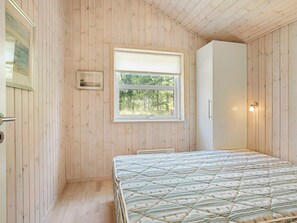 Ein Zimmer mit Holzwänden, einem Bett mit gesteppter Decke, einem Fenster mit Blick auf Bäume und einer Wandleuchte.