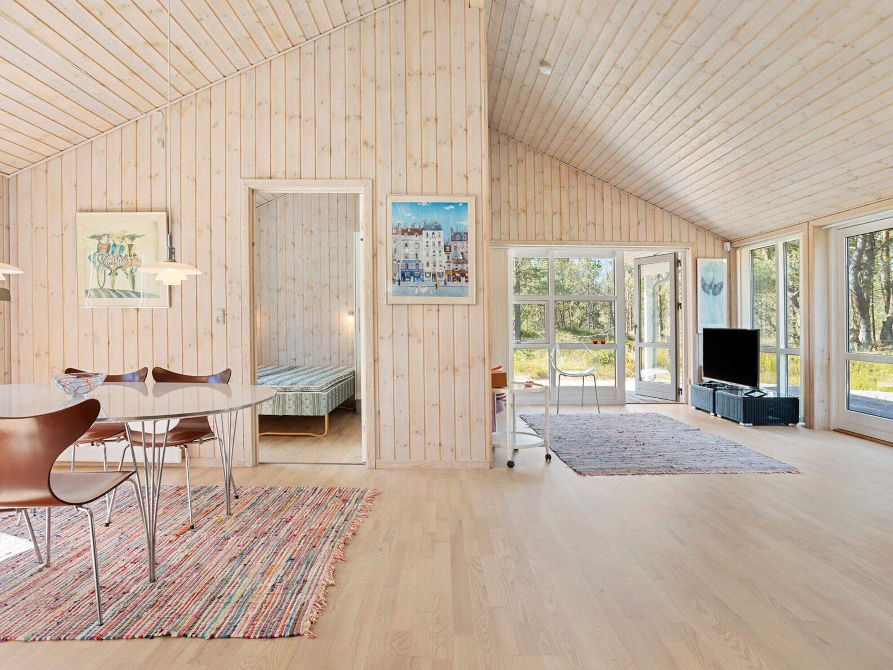 A spacious room with a high ceiling, wooden walls, a dining area with chairs and a table, a bed, a television, and large windows offering a view of the outdoors.