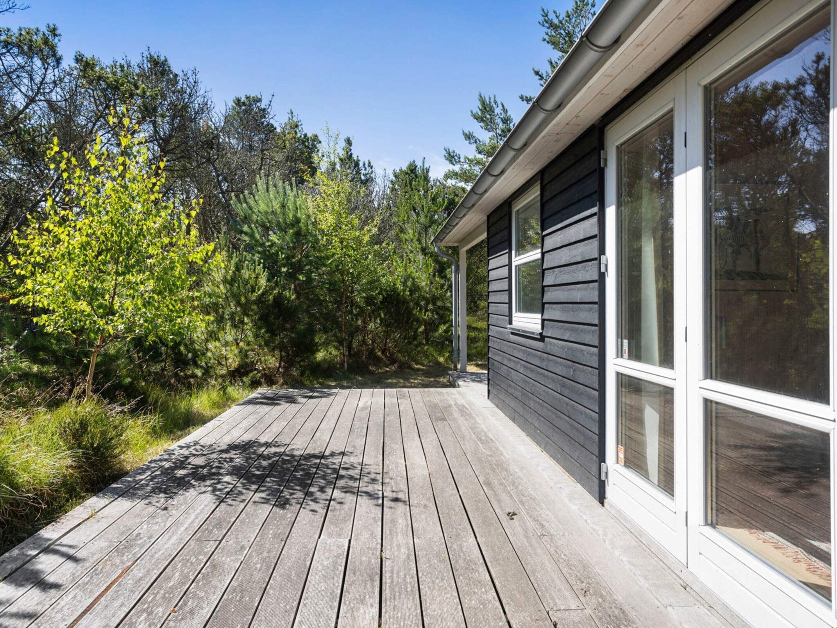 Een houten terras leidt naar een huis met grote ramen en een donkere gevel.