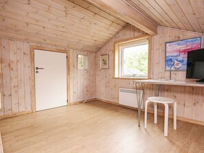 A room with wooden walls and ceiling, a white door, a window with a view, a desk with a computer, and a chair.