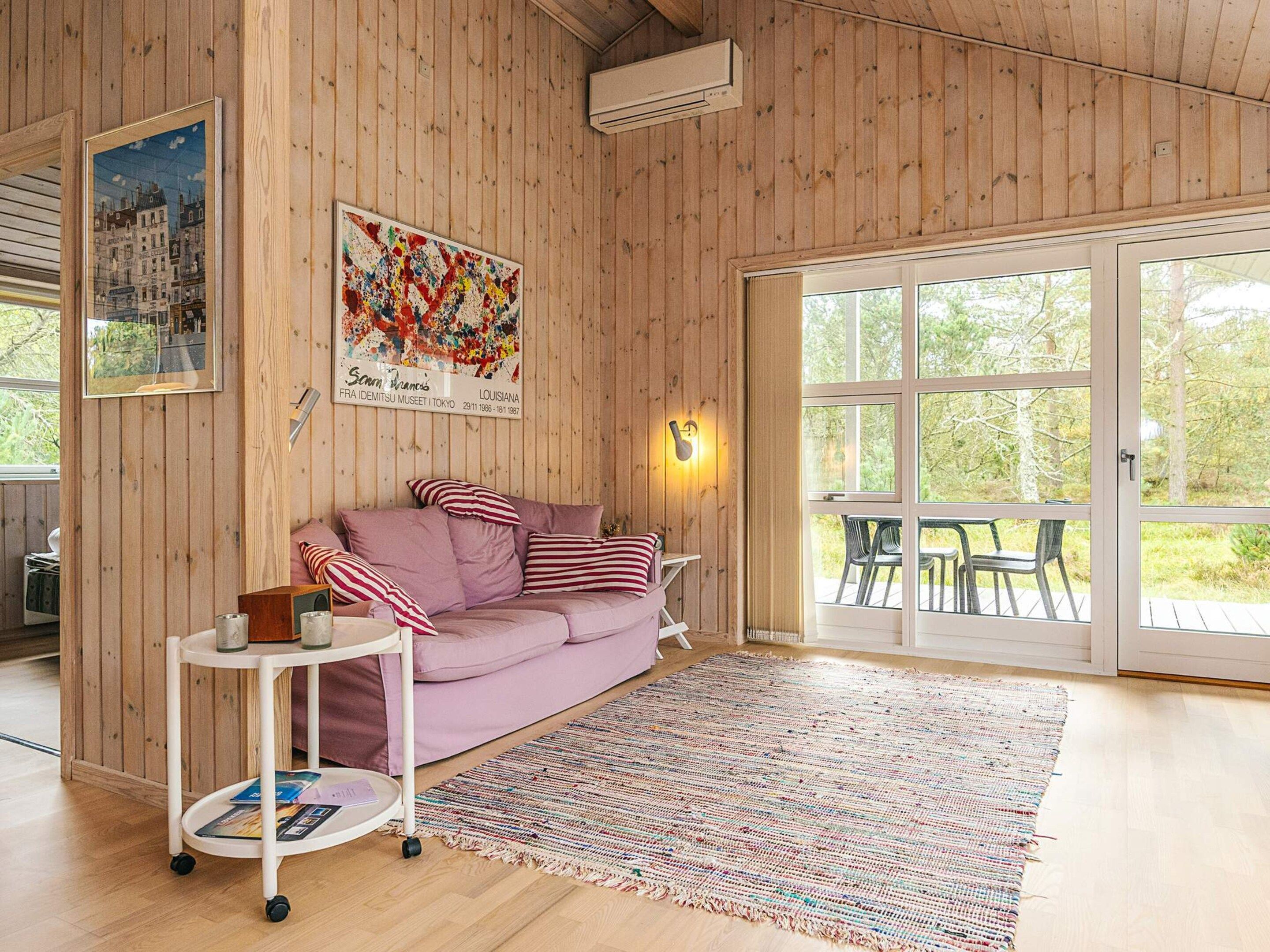 Ein Zimmer mit Holzvertäfelung, einem violetten Sofa, einem weißen Beistelltisch, einem farbenfrohen Teppich und einem großen Fenster mit Blick auf Grünflächen.