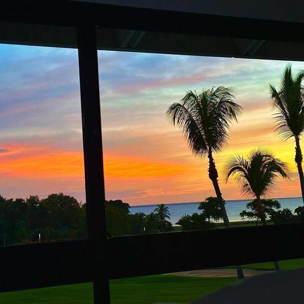 Sonnenuntergangblick durch ein Fenster mit Palmen und weiter Wasserfläche im Hintergrund.