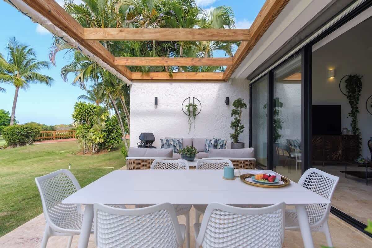 Ein möblierter Außenbereich mit weißem Tisch und Stühlen, einer Holzpergola und Blick auf einen üppigen Garten mit Palmen.